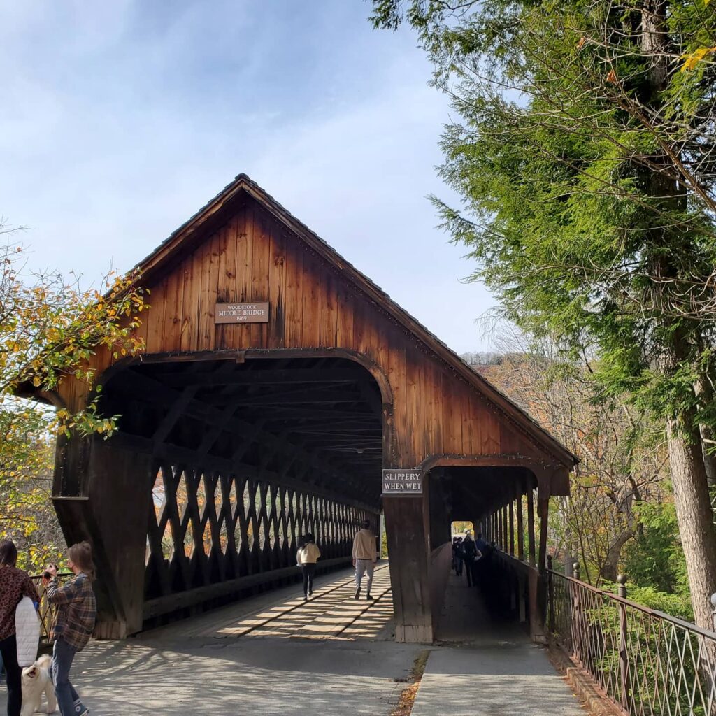 Wooden covered bridge with a peaked roof beside evergreen trees under a blue sky