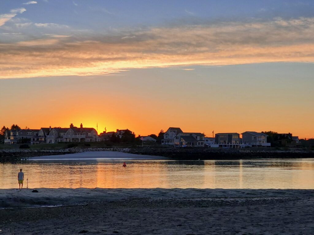 Sunset over calm water with houses along the shoreline and a person standing on the sand